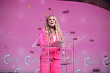 Woman with blonde hair stands at a podium wearing a pink suit.