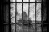 a shadowed figure is seen from behind bars as in jail.