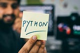 A guy holding a sticky note with Python written on it, while smiling in the background
