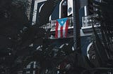 Dark, lush vegetation fills the bottom half of the image. In the background a Puerto Rican flag hangs from a second story balcony of a white building in San Juan.