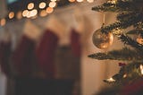 A gold, sparkly ornament hanging from a Christmas tree. Stockings and lights blurred in the background, over a fireplace.