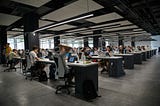 People sitting in rows of desks in an office, working on computers under fluorescent lighting.