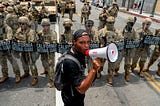 A young man wearing all black with a backpack and a megaphone stands before a line of California National Guard members in uniform with shields.