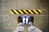 View down on feet standing on a “stand here” mark, behind a black and yellow warning line.