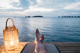 Next to a glowing lantern, a person’s legs crisscrossed on a mat on a dock overlooking a lake