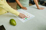 A person explaining a food chart to another individual and a pear kept on a table by the side.