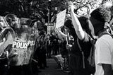 A black and white photo of police with shields facing protesters.