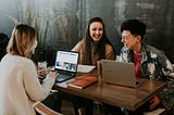 Three smiling women sit on a table with laptops in front of them