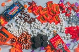 Spread out on a table there’s halloween candy and decorations with marshmellows, pretzels, oreos and raisonets sprinkled on the table too