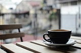 A coffee cup and saucer on a wooden table. A wooden can be seen in the background, and there are some buildings in the background, but the background is blurred.
