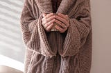 Wrapped in a cozy, thick brown knit sweater, a woman holds a steaming mug in her hands.