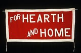 A handmade banner set against a black background. The banner is red with white lettering and white trim. It reads “For Hearth and Home.” It is believed to be a suffragette banner.