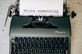 A typewriter with a piece of paper. On it: “Write something”