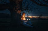 A young man sitting by a tree at night and reading a book