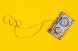 A yellow background with a clear cassette tape with the tape pulled out and unwound slightly.
