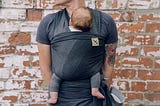 A father is holding his son in a baby sling attached to the front of his body. The father has colourful tattoos on his right arm. Baby has dark hair and bare feet.
