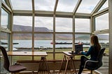 woman on a laptop looking through the window at a boat dock from inside a warm coffee shop