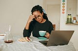 Woman at laptop with coffee, finger on forehead