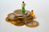 Two very small figurines standing and sitting on a pile of coins, looking in opposite directions.
