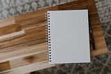 A wooden tabletop with a spiral bound notebook. Next to the notebook is a pen.