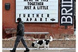 A man and dog walk past a sign that reads, “We’re in this together. Just a little further apart.”