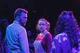 Three performers on stage, bathed in purple light. Two of them look suspiciously at a third, whose back is to us.