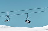 Ski lift chairs against a clear blue sky, with two skiers on the further chair above a sparkling snow-covered slope. The descending chair is empty.
