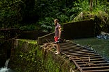 A white bearded man in red shorts and a tank top stands on top of a rock dam covered with moss. There is a wide wooden ladder lying sideways on top of the dam.