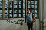 Young man wearing sunglasses, a red hoodie with design partially hidden by a denim vest, black jeans, and a backpack. He is standing next to a wall with a large window full of promotional posters. Beneath the wall is spray-painted 666 and random letters.