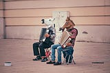 Two men playing a musical instrument on the street with horse masks on their faces.