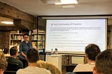 Me standing at a podium in front of a big screen at the Made Tech offices, pulling a funny face and introducing our Community of Practice