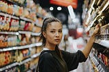 A woman with light skin and dark hair in a pontytail lifting her arm to pick up an item from the shelf at the grocery store.