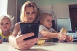 Image of a woman sitting at a table, staring at her cell phone with a small child on either side of her