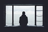 A person sits on the edge of a stone window opening, flanked by vertical metal bars on either side. They look out onto an ocean view. Clouds are gathering in the distance.