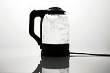 A transparent kettle with black plastic handle, lid and base, is filled with boiling water.