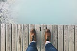 Person looking down at their shoes as they stand on wooden walkway over cloudy water.