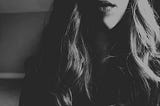 Black and white photo of the bottom half of a woman’s face from the top of her lips to just below her shoulders. Her blonde wavy hair falls over her shoulders and she’s wearing lipstick. She’s not smiling.