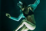 Underwater. Model poses in a dancer-like position with arms outreached and legs in a zigzag shape. Bubbles erupt from her mouth, slightly obscuring her serene expression.