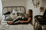 A person sitting in their bed with a cup of coffee or tea. They look tired.