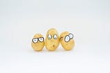 Three small potatoes lined up in front of the camera. They are in the center of the image and each one is wearing a paper cut out with fake eyes stickered onto the top half of the potato. The first one is wearing glasses and their eyes are shut the second looks surprised and the third looks confused