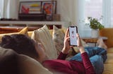 a person lying on sofa holding a smartphone with a person’s profile on screen