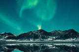 a green aurora dances across a starry sky, above a mountain, reflected in a perfectly smooth lake
