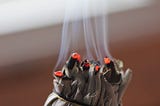 A close up photograph of a sage bundle, lit at one end with smoke rising from the ignited leaves.