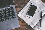 A laptop, diary, and smartphone are laid on a rustic wooden table. There’s a checklist in the diary, parts of it are complete