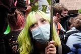 Photo of a protester with yellow hair and a mask carrying an olive branch in a crowd