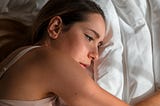 Young dark-haired woman, with pensive expression, in bed on white sheets