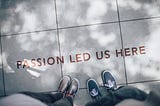Shoes and legs of two people are visible with text printed on floor “Passion Led Us Here”
