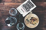 A book about advice sits on a wooden table with a carafe, glass, and the remains of a meal on a plate.