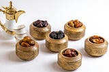 An image of a bunch of copper bowls with different a variety of dates in each, along with a stack small white tea-cups and an ornate tea pot with polished gold colored accents and handle.