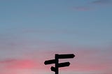 A signpost stands silhouetted against the sky at sunset.